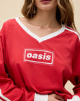 A woman with wavy brown hair wears the DayDreamer Oasis Box Logo red long-sleeve shirt featuring white trim and "Oasis" in white text inside a red box, standing against a beige background.