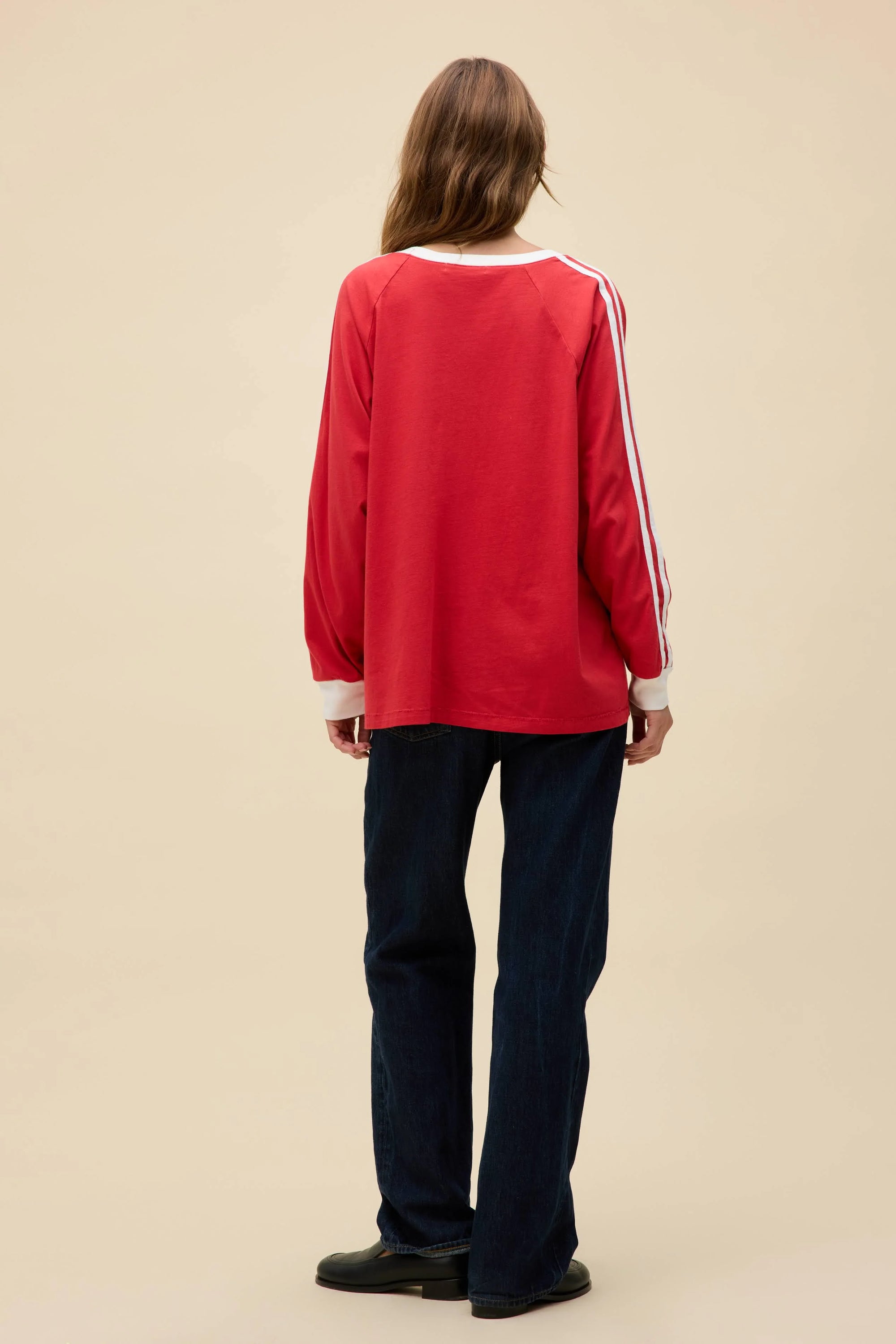 A person with long hair faces away from the camera, wearing the DayDreamer Oasis Box Logo red long-sleeve top with white sleeve stripes, dark jeans, and black shoes against a plain beige background, evoking Britpop style.