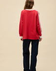 A person with long hair faces away from the camera, wearing the DayDreamer Oasis Box Logo red long-sleeve top with white sleeve stripes, dark jeans, and black shoes against a plain beige background, evoking Britpop style.