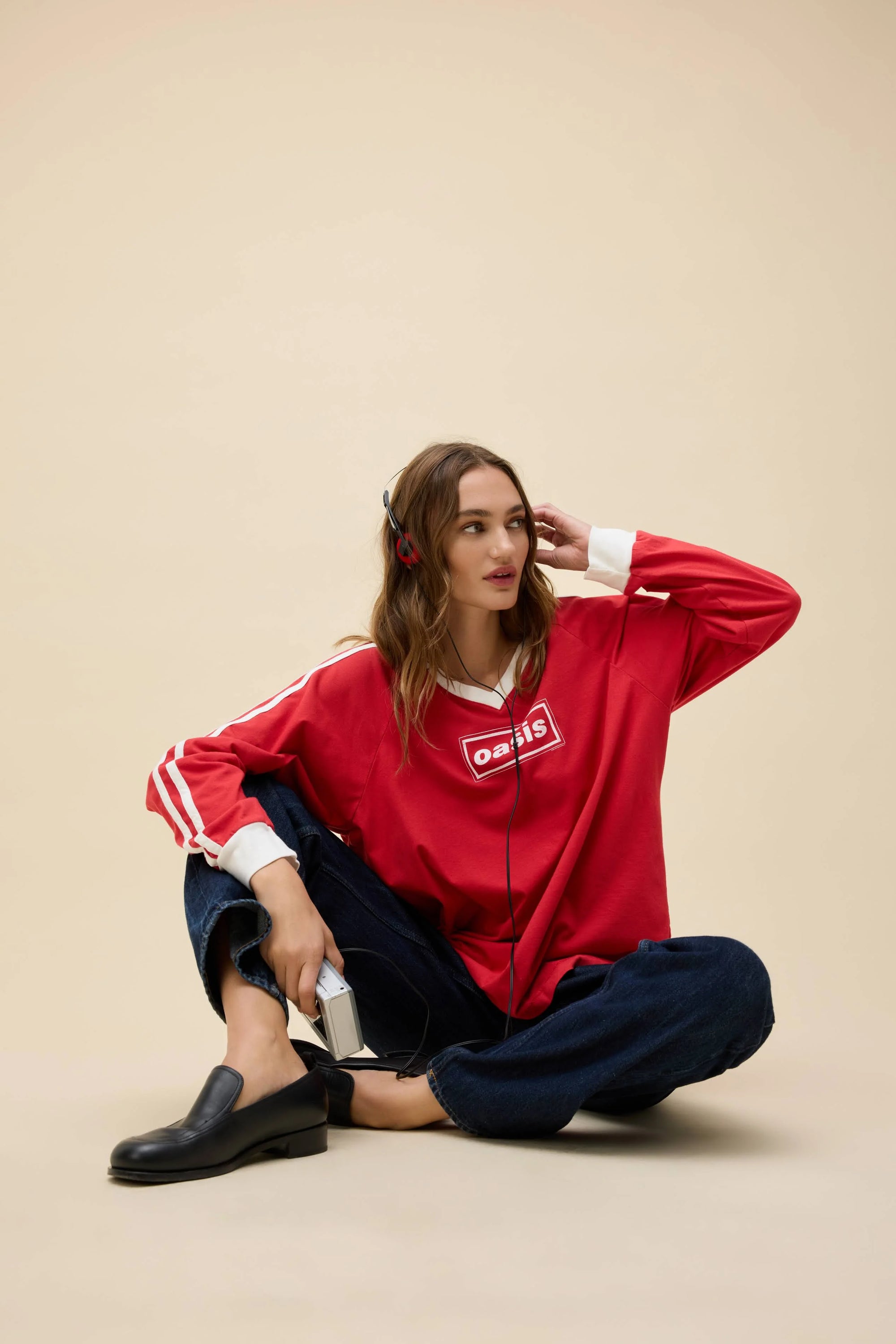 A woman in the DayDreamer Oasis Box Logo sweatshirt sits cross-legged on the floor, wearing headphones and holding a cassette player, lost in Britpop vibes, against a plain beige background.