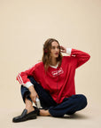A woman in the DayDreamer Oasis Box Logo sweatshirt sits cross-legged on the floor, wearing headphones and holding a cassette player, lost in Britpop vibes, against a plain beige background.