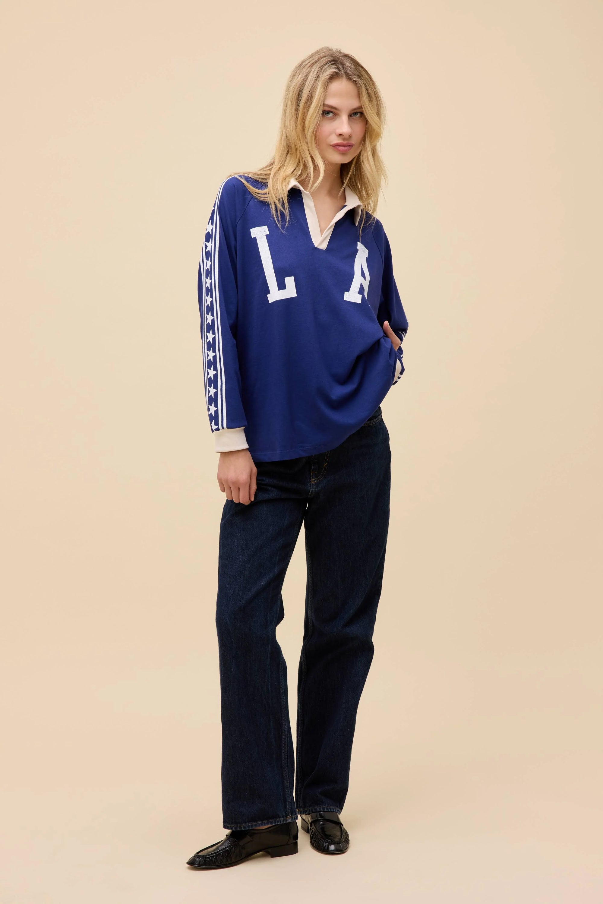 A blonde woman wears the DayDreamer Los Angeles Stars Rugby, an oversized blue long sleeve shirt with "L A" on the front, paired with dark jeans. She stands against a plain beige background, looking forward with a slight smile.