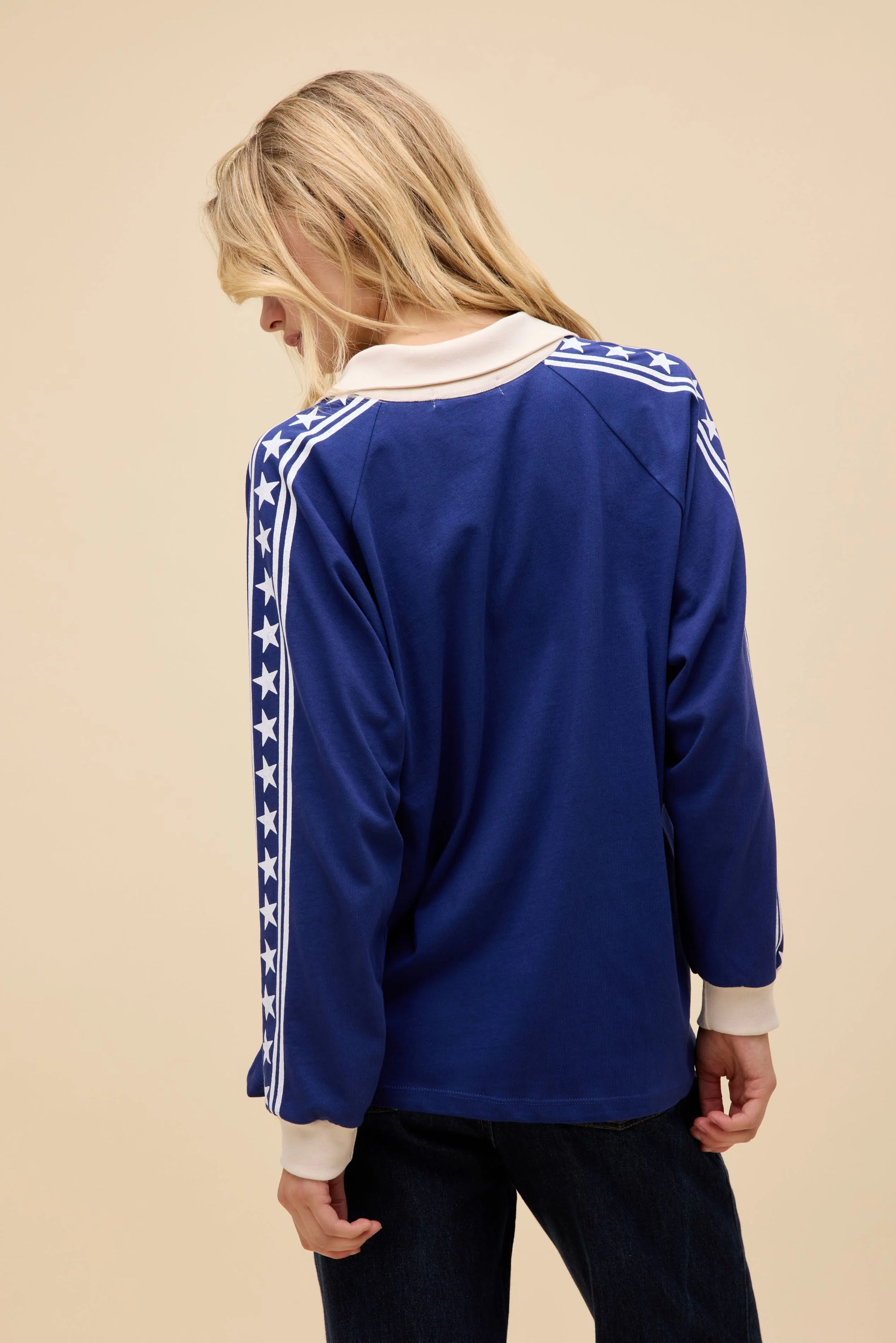 A person with long blonde hair faces away, wearing the DayDreamer Los Angeles Stars Rugby top—featuring white cuffs, collar, and star-striped sleeves—against a plain beige background.