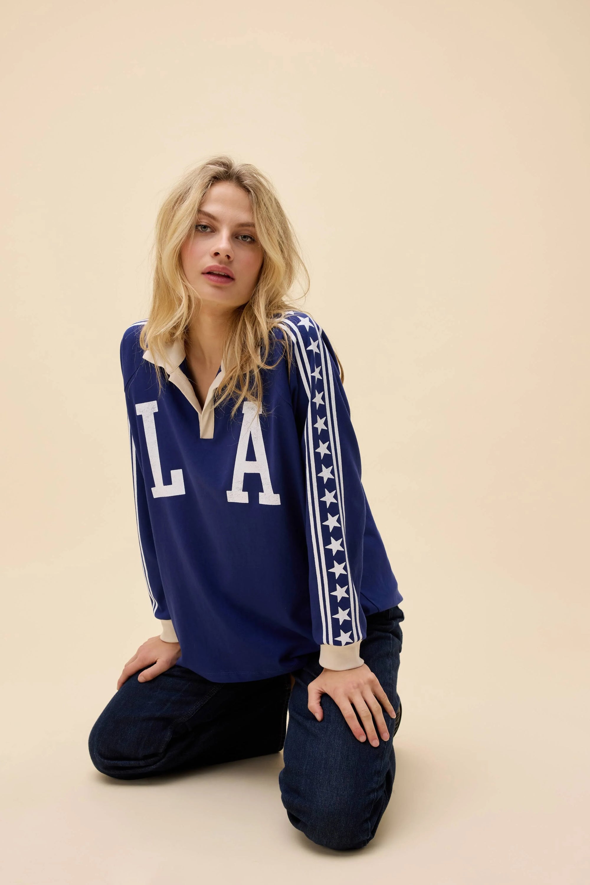 A blonde woman kneels on the floor against a plain beige background wearing the DayDreamer Los Angeles Stars Rugby long sleeve shirt with "LA" and star-striped sleeves.