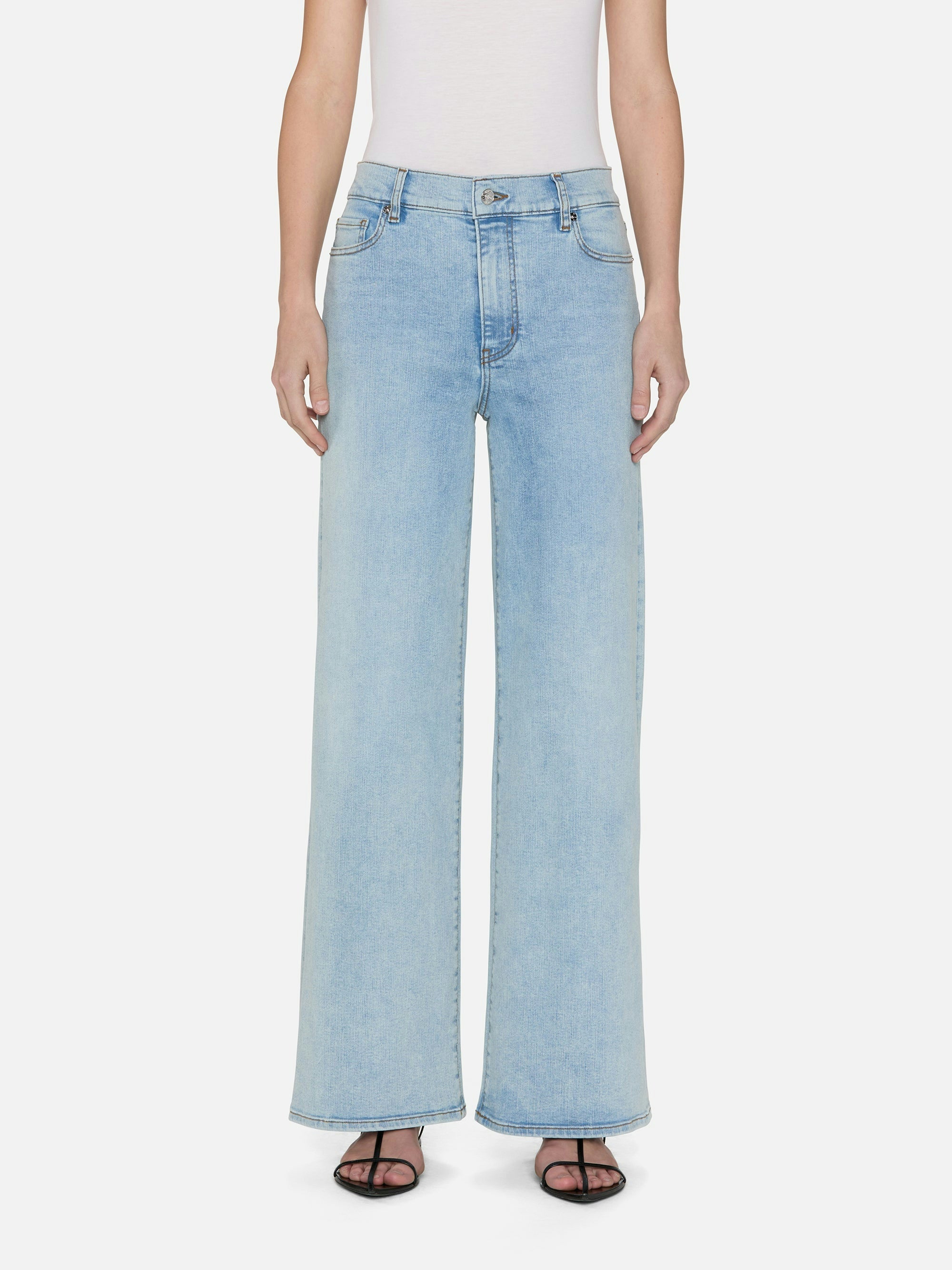 A person is wearing FRAME's Le Slim Palazzo light blue wide-leg jeans, crafted from sustainable cotton in a chic palazzo silhouette with a high rise and two front pockets. They wear a white sleeveless top and black open-toe sandals against a white background.