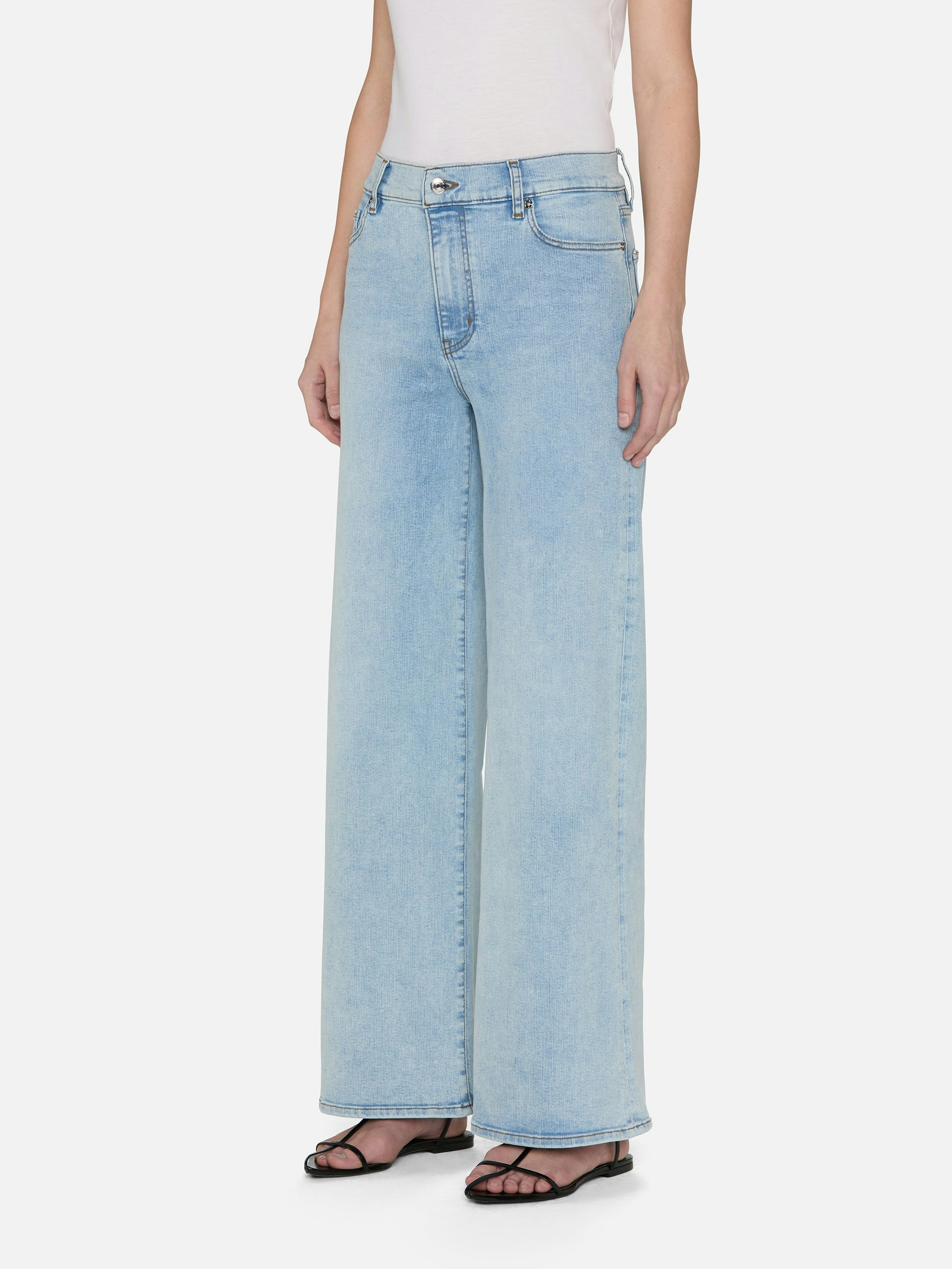 A person wears FRAME's Le Slim Palazzo light blue wide-leg jeans made from a sustainable cotton blend paired with a white top, standing casually. These high-rise jeans with pockets are stylishly completed with black sandals.