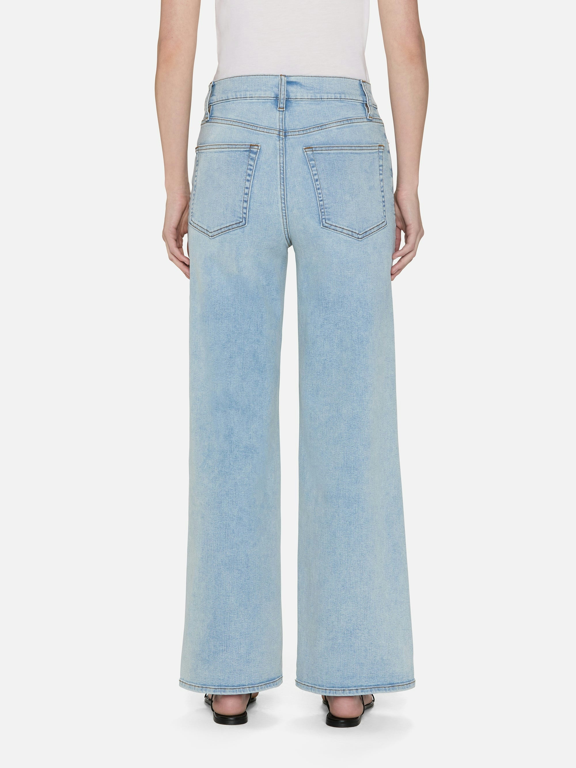 A person stands against a white background in FRAME's Le Slim Palazzo jeans, featuring a high-rise fit, light blue hue, sustainable cotton blend, wide-leg palazzo silhouette, and two back pockets. They pair the jeans with black heels for an effortlessly chic look.