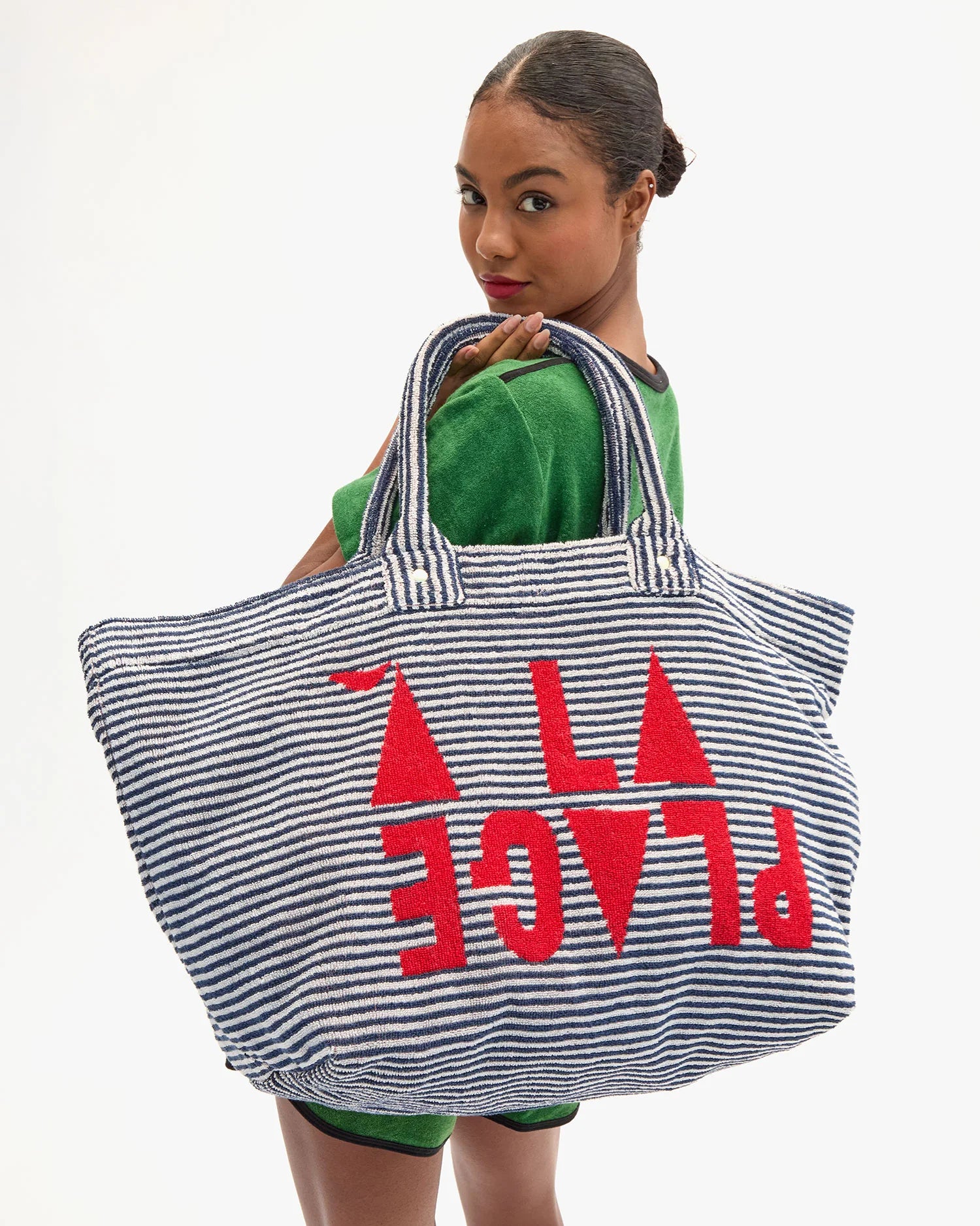 A woman in a green outfit holds the Clare Vivier La Plage Tote, a large blue and white striped beach bag with bold red “A PLACE” lettering, while glancing back at the camera against a white background.