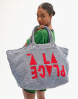 A woman in a green outfit holds the Clare Vivier La Plage Tote, a large blue and white striped beach bag with bold red “A PLACE” lettering, while glancing back at the camera against a white background.