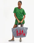 A woman in a green short-sleeve top and matching shorts holds the Clare Vivier La Plage Tote, a large blue-and-white striped cotton terry bag with red "A LA PLAGE" text. She wears striped slip-ons, standing against a plain white background.