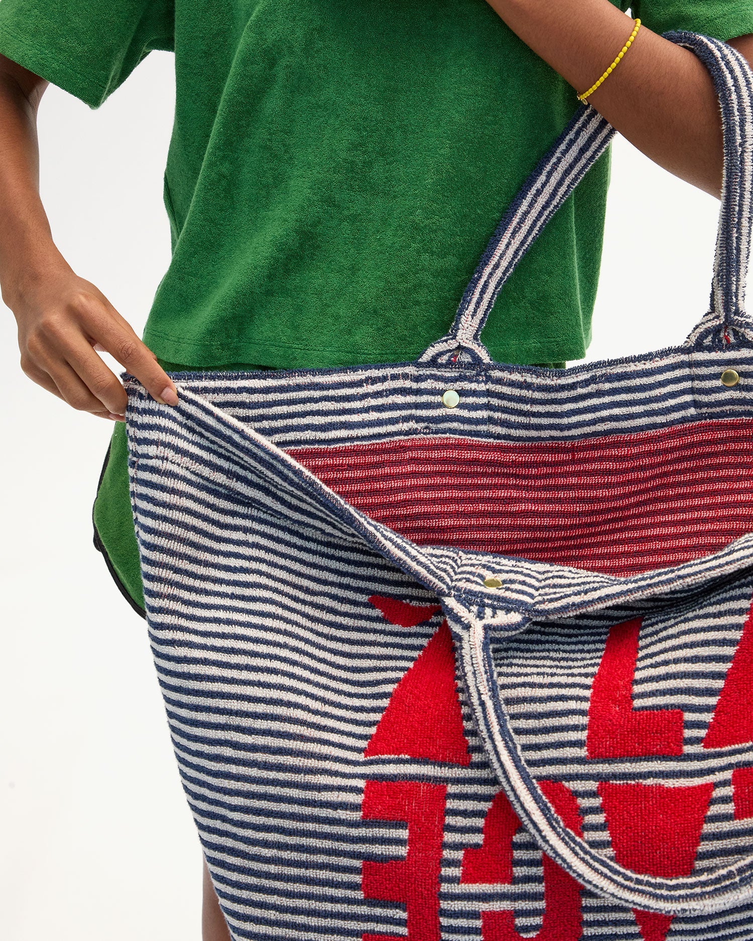 A person in a green shirt holds open the Clare Vivier La Plage Tote, a large blue and white striped cotton terry beach bag with red text inside. One visible arm wears a yellow bracelet; background is plain white.