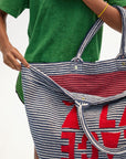 A person in a green shirt holds open the Clare Vivier La Plage Tote, a large blue and white striped cotton terry beach bag with red text inside. One visible arm wears a yellow bracelet; background is plain white.