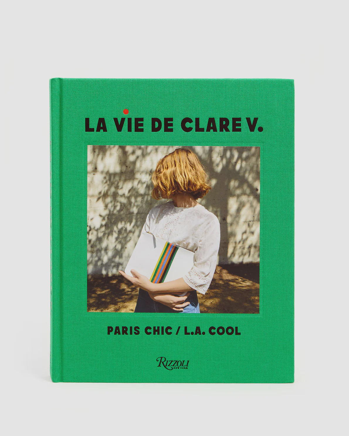 The image features a book with a green cover titled "La Vie De Clare V." by the brand Clare Vivier. A woman with blonde hair, dressed in a white lace top, holds a stylish clutch. The background is illuminated with dappled sunlight on the wall, emphasizing the subtext: "Paris Chic / L.A. Cool.