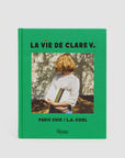 The image features a book with a green cover titled "La Vie De Clare V." by the brand Clare Vivier. A woman with blonde hair, dressed in a white lace top, holds a stylish clutch. The background is illuminated with dappled sunlight on the wall, emphasizing the subtext: "Paris Chic / L.A. Cool.