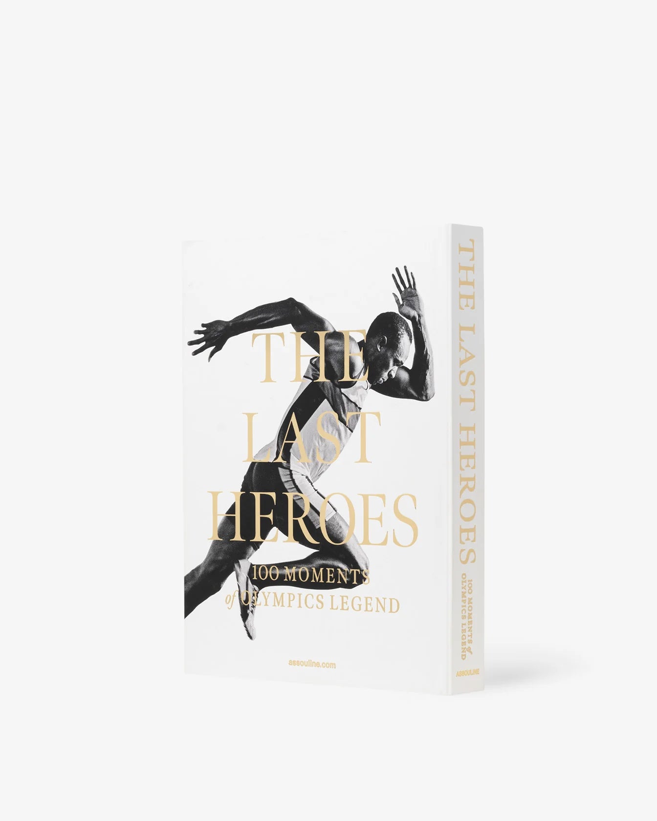 The image features an Assouline book titled "The Last Heroes: 100 Moments of Olympics Legend," with a black and white photo of an athlete in mid-sprint on the cover. The book, which delves into Summer Olympics history, is positioned standing upright, displaying both the front cover and spine.