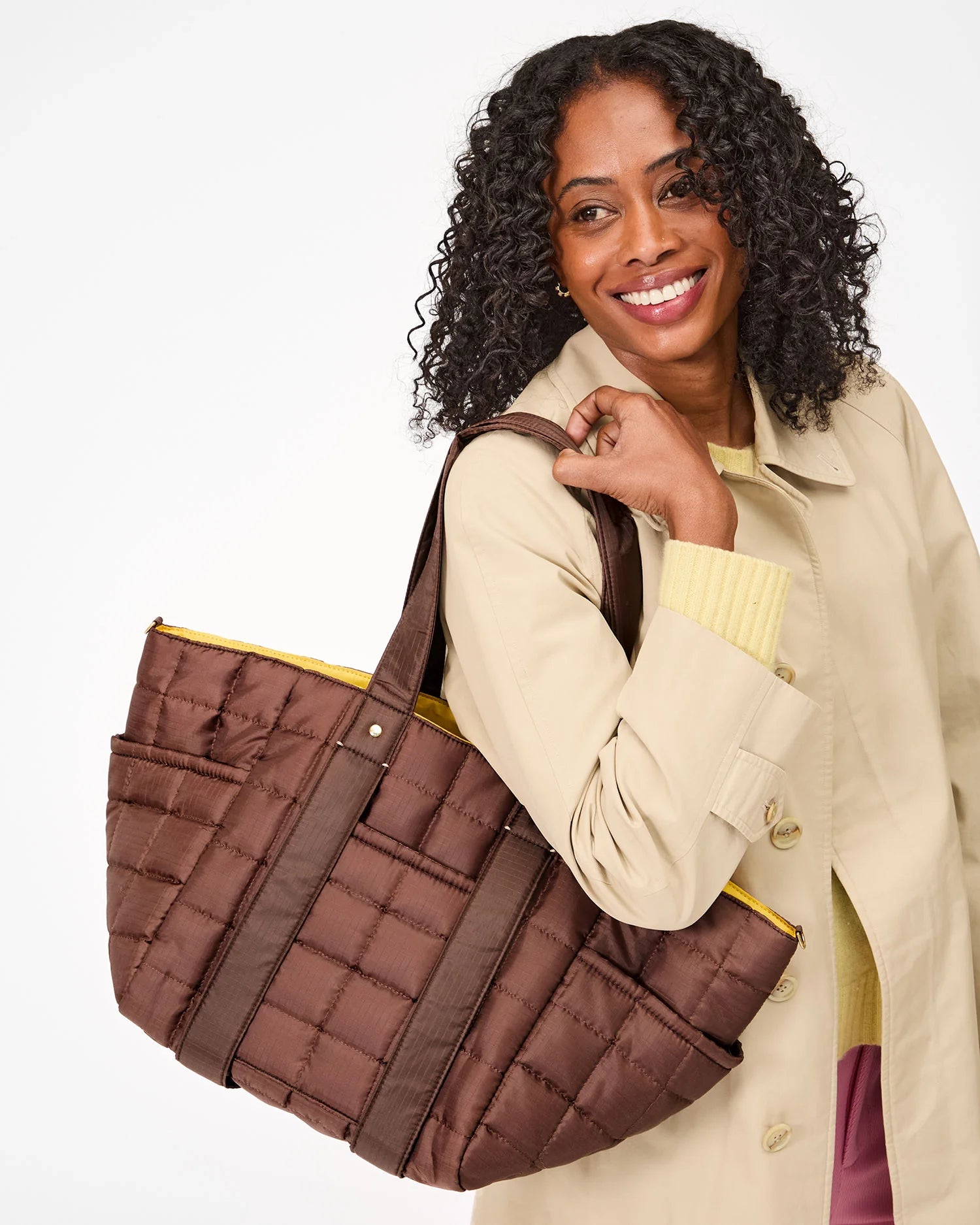 A woman with curly hair smiles, shoulder carrying the large quilted Le Box Tote Sportif by Clare Vivier. She's wearing a light beige coat and a yellow top against a plain white background.