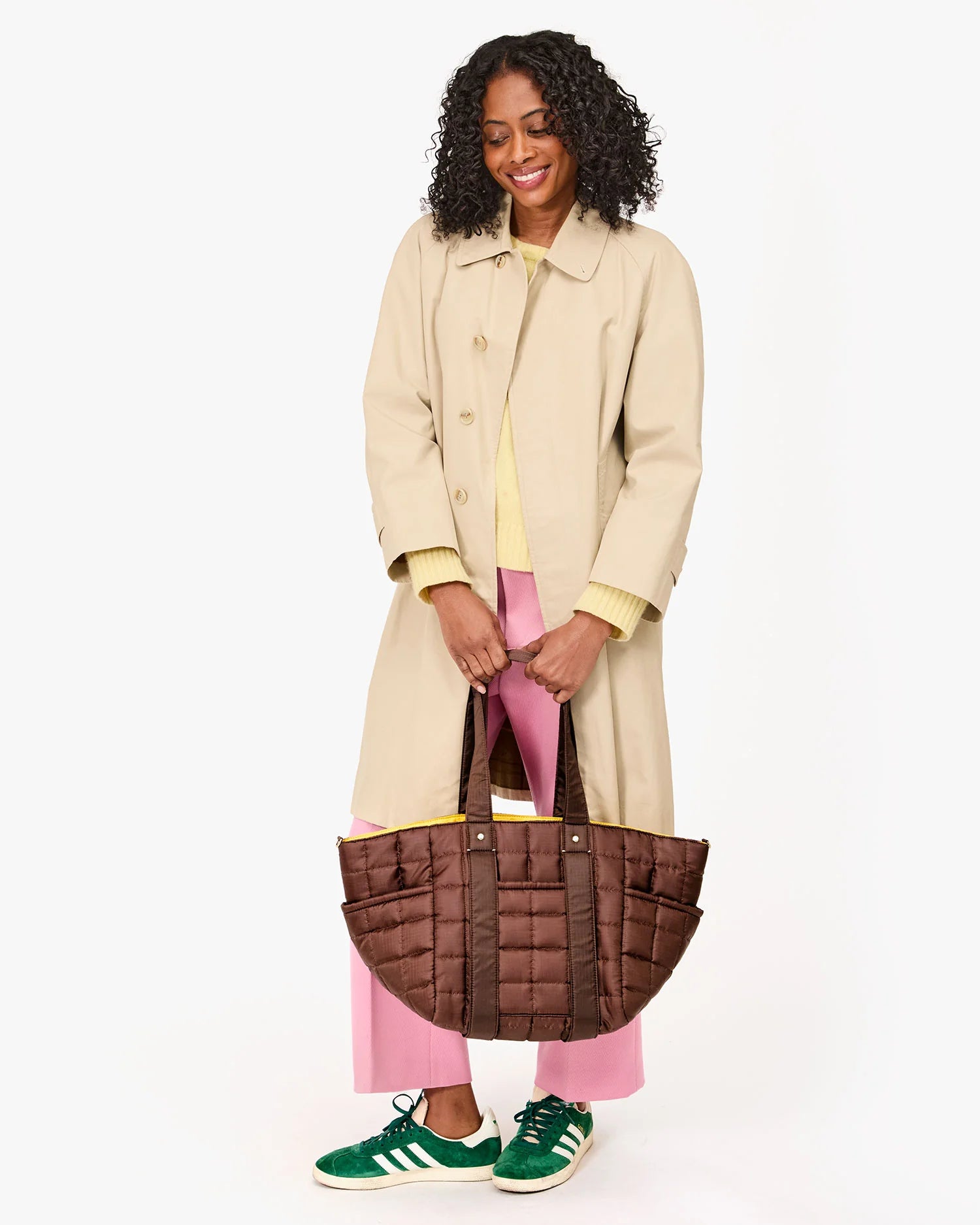 A curly-haired person in a beige coat, yellow top, and pink pants carries Clare Vivier's Le Box Tote Sportif. They're smiling while wearing green and white sneakers. The tote's lightweight nylon fabric makes it perfect for easy shoulder carry.