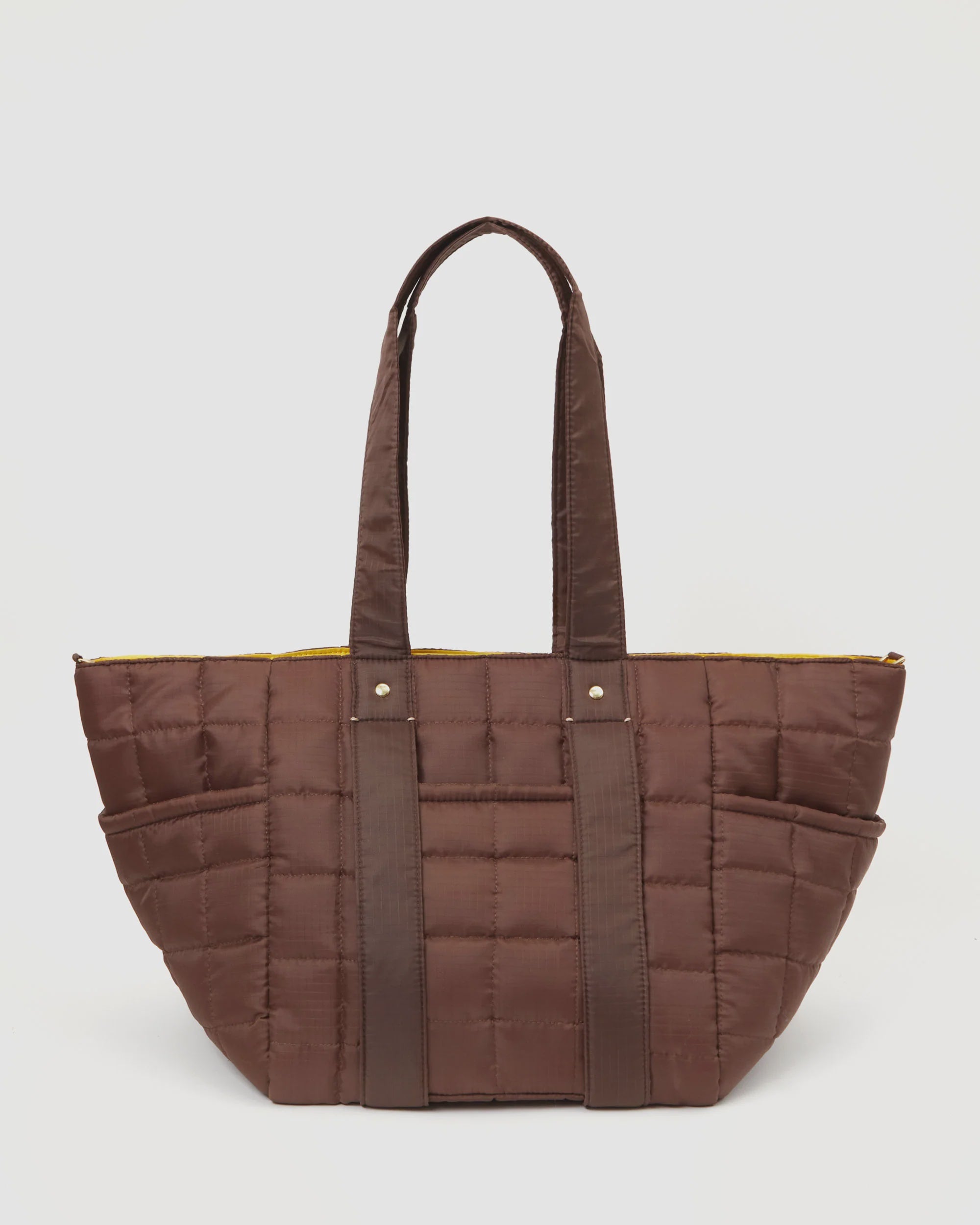 The Clare Vivier Le Box Tote Sportif is a stylish brown quilted bag with long straps and a U-shaped design, featuring meticulous checkered stitching. Its interior is lined with bright yellow fabric and crafted from vegan materials for an eco-conscious touch.