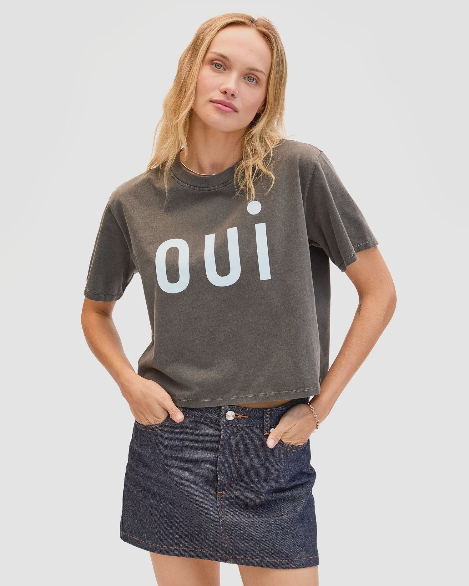 A woman with long blonde hair wears the Clare Vivier Le Carre Tee, a dark gray 100% cotton boxy shirt with "oui" in large white letters, paired with a dark denim skirt. She stands against a plain light background, hand in her pocket.
