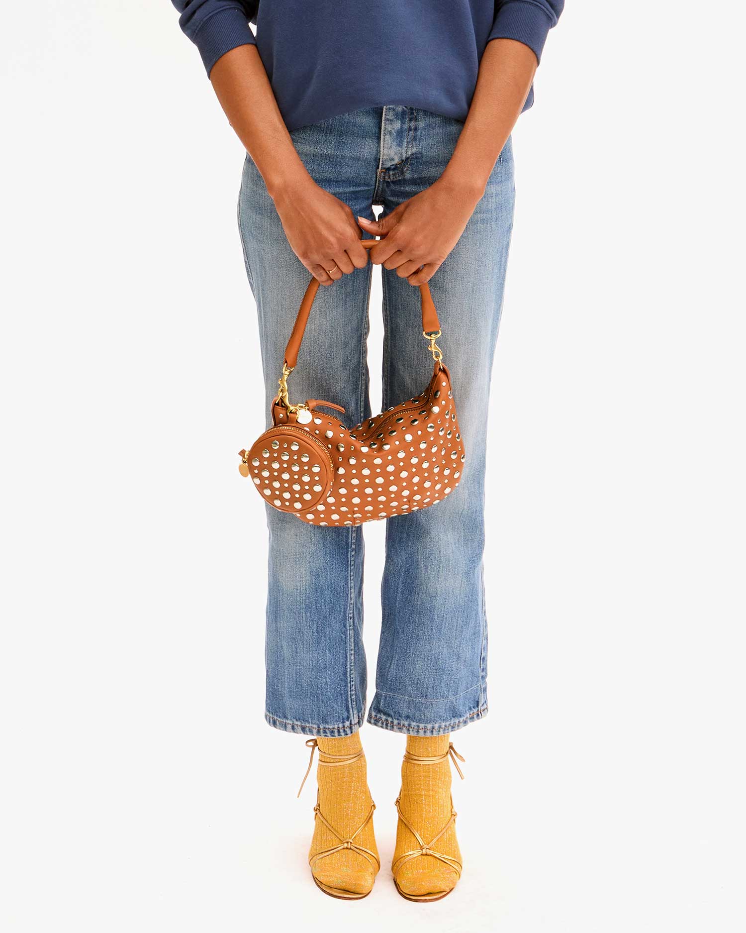 A person in a blue sweater and light blue jeans holds Clare Vivier's brown polka dot handbag with a chic zipper. They complete their ensemble with yellow socks and the stylish Le Disco yellow woven sandals, adding a touch of flair against the plain white background.