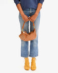 A person wearing a blue sweater and light blue jeans is holding the Clare Vivier "Petite Moyen" brown handbag, which is adorned with chrome studs and white polka dots, featuring a sleek zipper closure. They are styled with yellow socks and mustard yellow heels, standing against a white backdrop.