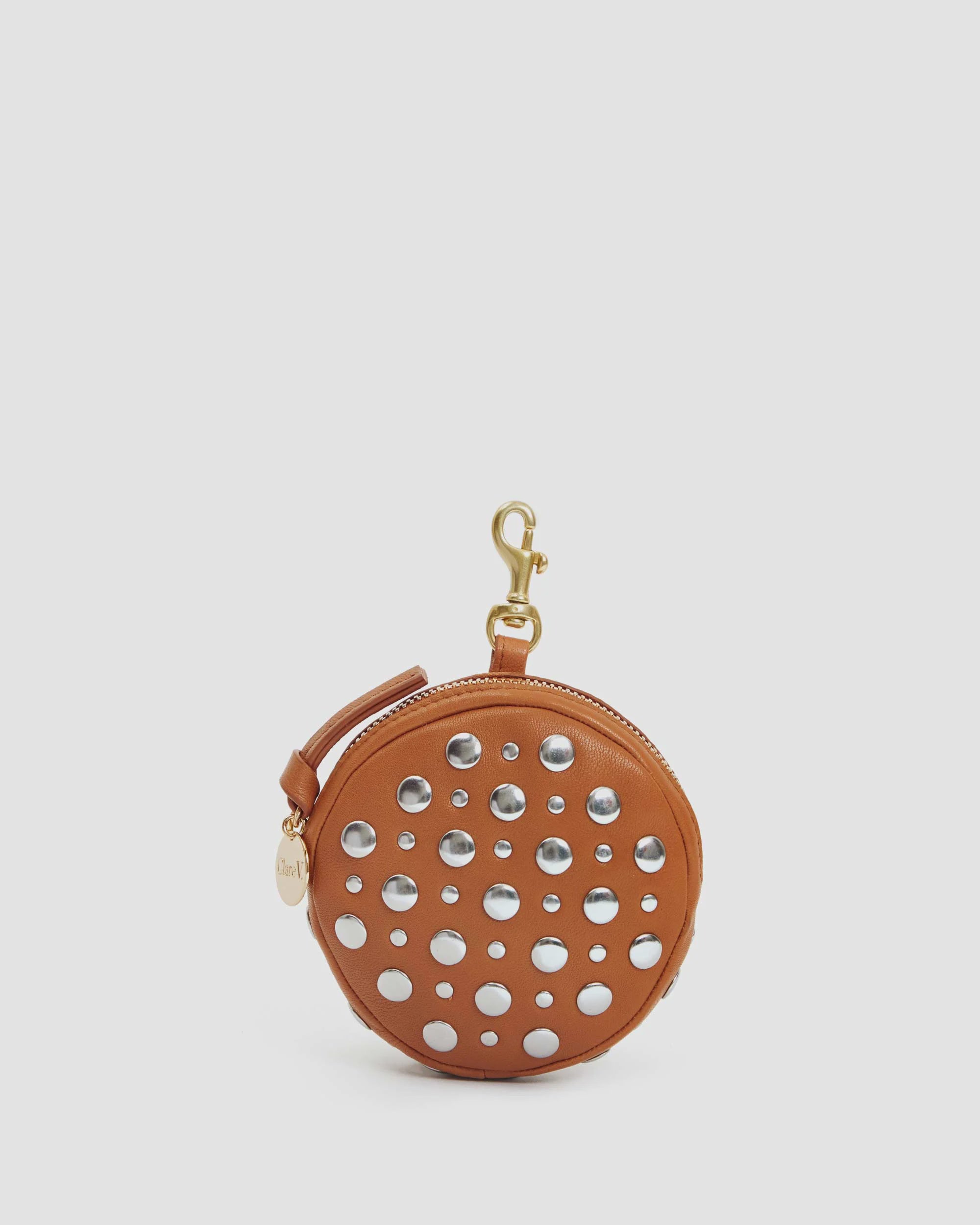 The chic "Le Disco" pouch by Clare Vivier is a round, tan studded nappa leather accessory adorned with silver studs in a polka dot pattern. It features a gold zipper and clasp, along with a small leather tag, all set against a plain light gray background. Made in India, it truly stands out as a stylish piece.