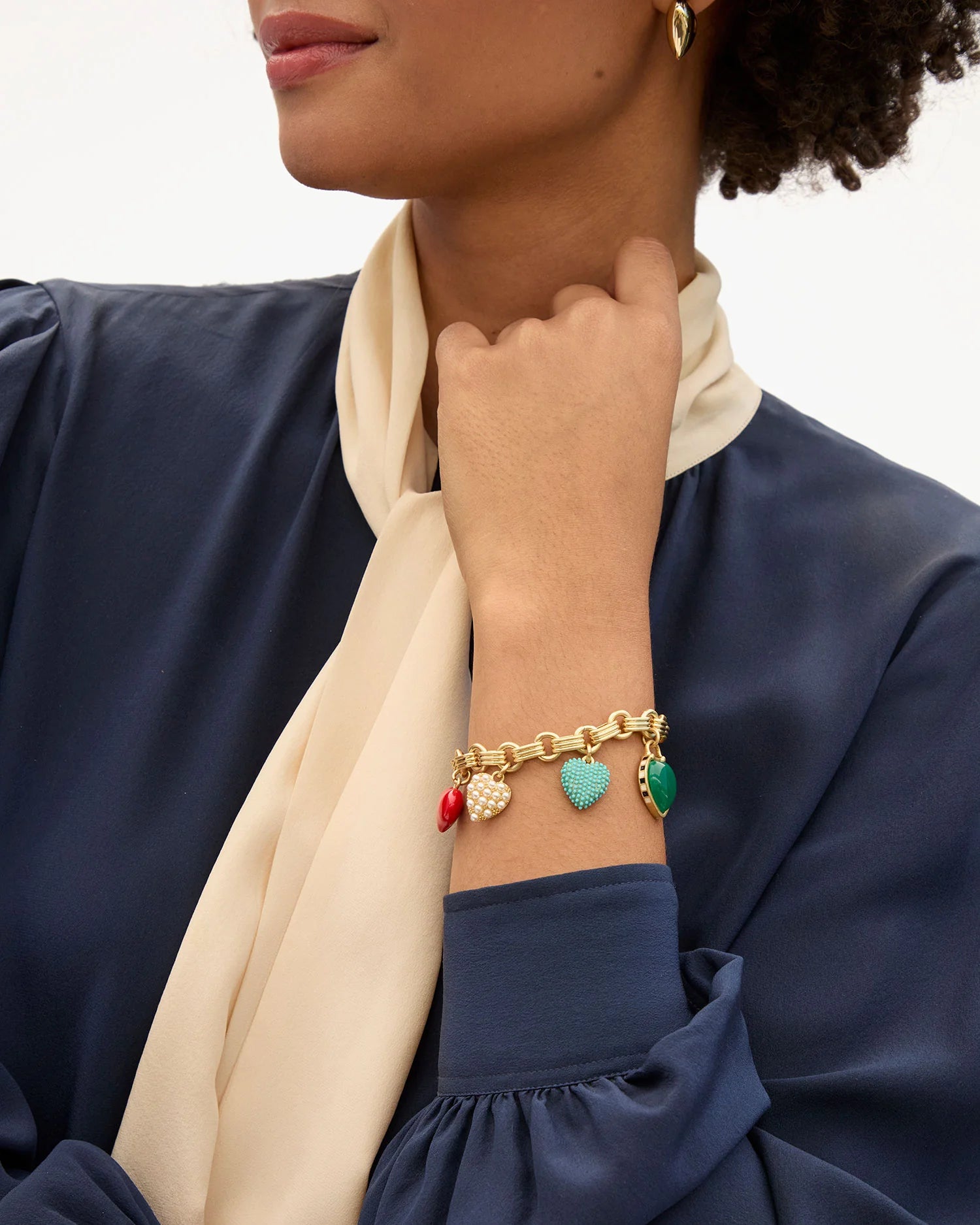 A woman in a navy blouse and cream scarf displays the Clare Vivier LE Heartbreaker Bracelet, a gold-plated brass piece with red, turquoise, gold, and green heart charms, paired with matching gold earrings.