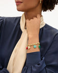 A woman in a navy blouse and cream scarf displays the Clare Vivier LE Heartbreaker Bracelet, a gold-plated brass piece with red, turquoise, gold, and green heart charms, paired with matching gold earrings.