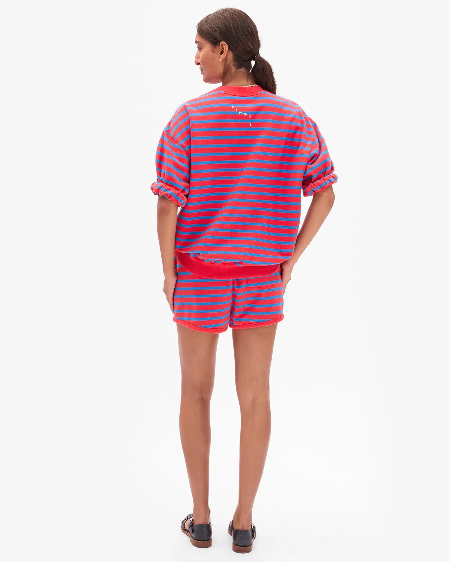 A woman with brown hair tied back wears the Clare Vivier Le Jogging Short—a red and blue striped cotton sweat short with an elastic waistband—paired with a matching short-sleeve sweatshirt and black sandals, set against a plain white background.