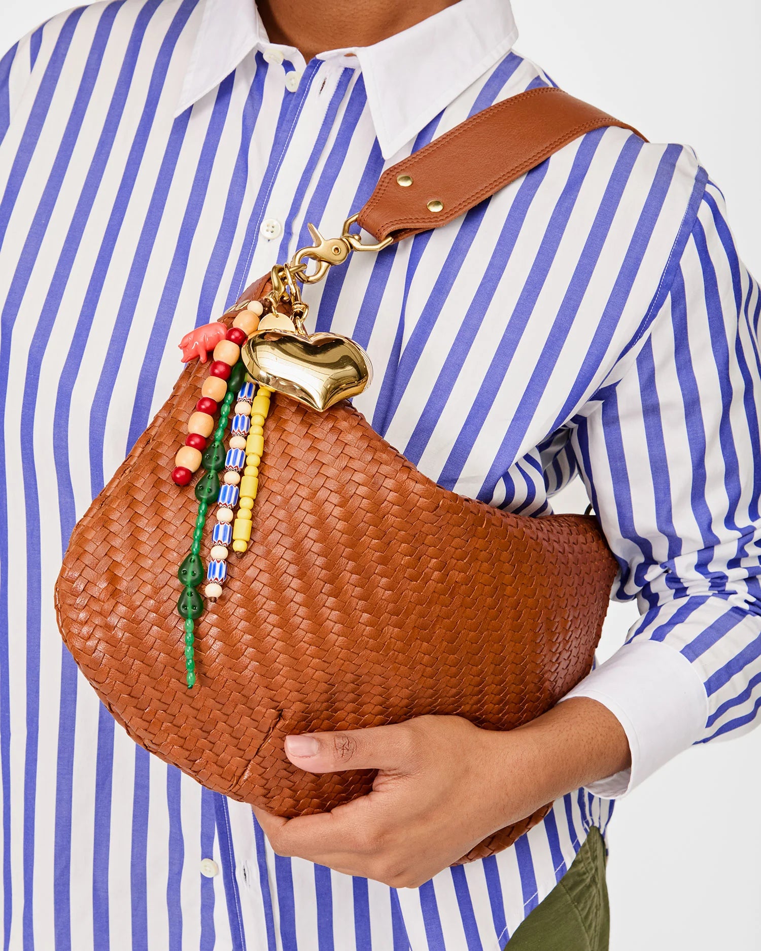 A person holds a Clare Vivier brown woven handbag with a wide strap, decorated with the Le Long Beaded Tassel and a vintage brass heart keychain, while wearing a blue and white striped shirt.