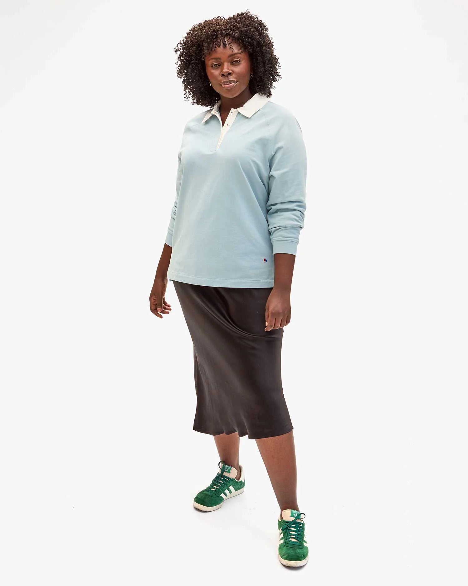 Wearing Clare Vivier's "Le Rugby," a long-sleeve rugby shirt in light blue with elbow patches, paired with a black skirt and green sneakers, a person with curly hair strikes a stylish pose against a white backdrop. Oui To Le Rugby!.