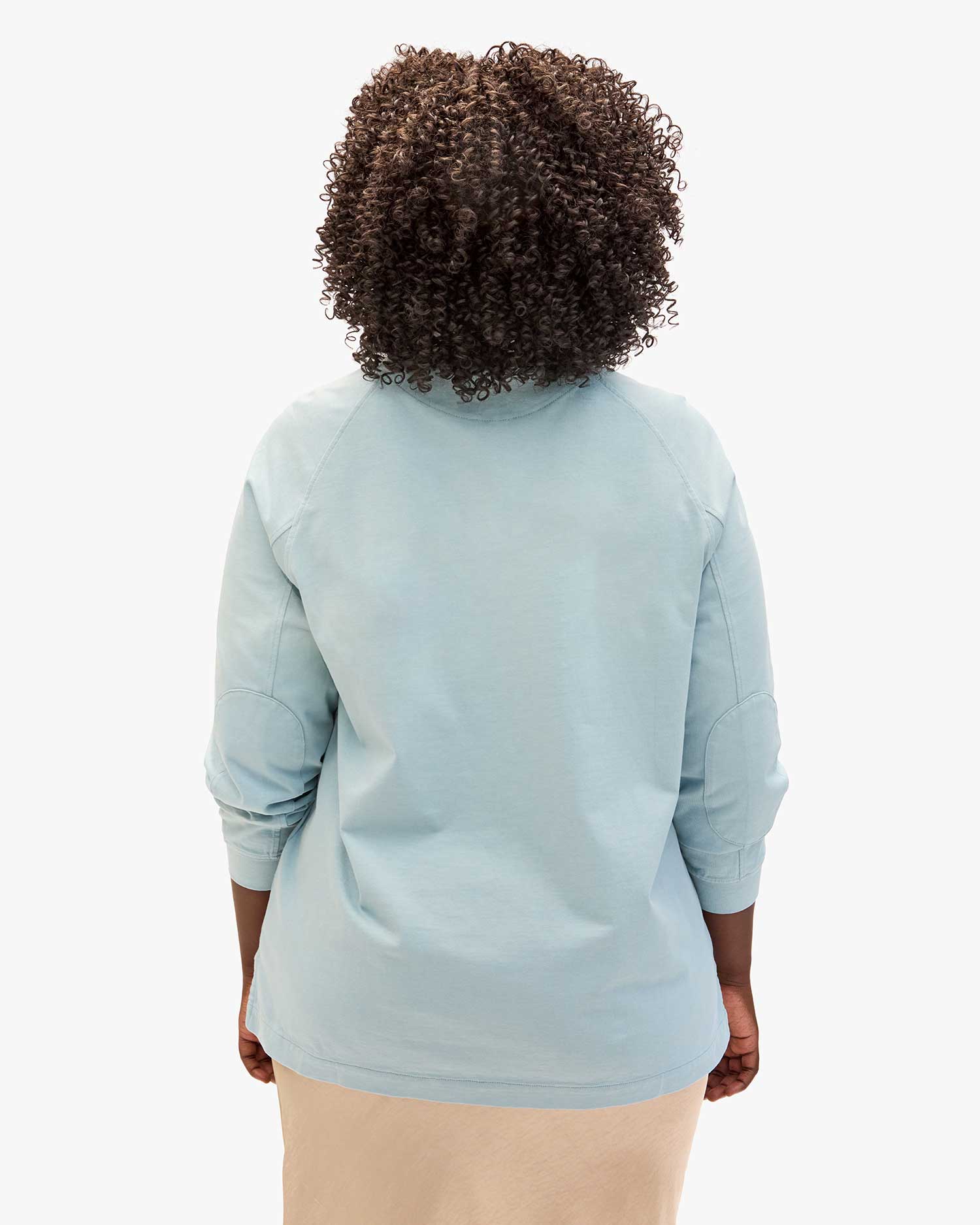 A person with curly hair is wearing the Le Rugby light blue long-sleeve shirt by Clare Vivier, featuring elbow patches, paired with a beige skirt, standing with their back to the camera against a plain white background.