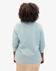 A person with curly hair is wearing the Le Rugby light blue long-sleeve shirt by Clare Vivier, featuring elbow patches, paired with a beige skirt, standing with their back to the camera against a plain white background.