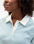 A person with curly hair wears Clare Vivier's "Le Rugby," a light blue heavyweight cotton collared shirt with a white collar and one signature red button. A heart-shaped earring adds flair, while the plain background complements their style.