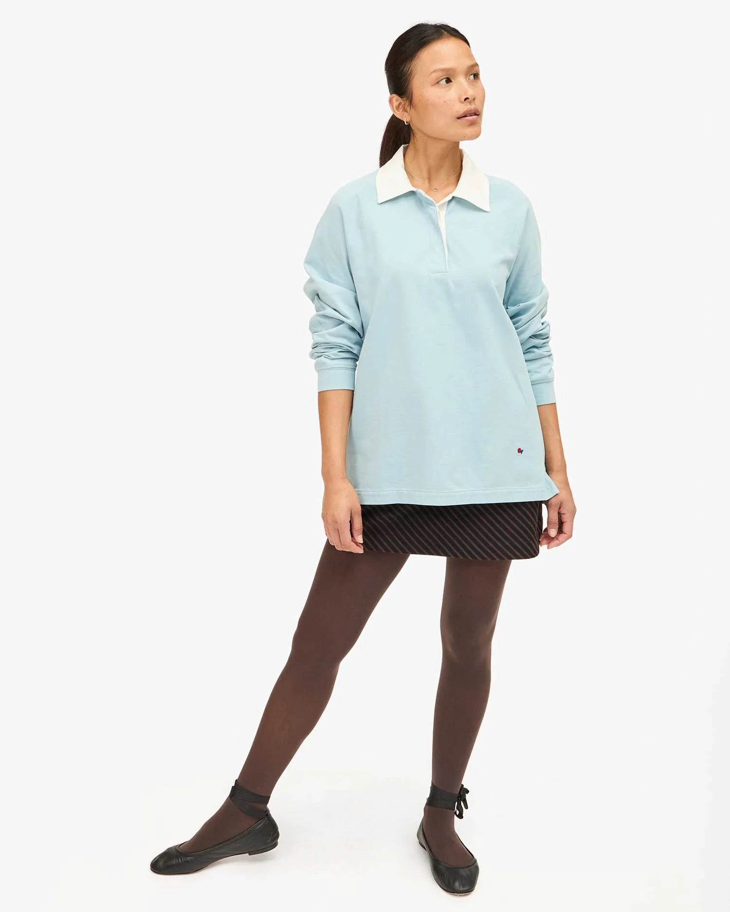 Against a white background, a person wears Clare Vivier's Le Rugby—a light blue long-sleeve top with a white collar and discreet elbow patches—paired with a black skirt, dark tights, and black shoes. Hair pulled back, they gaze to the side.