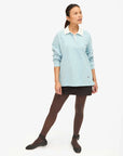 Against a white background, a person wears Clare Vivier's Le Rugby—a light blue long-sleeve top with a white collar and discreet elbow patches—paired with a black skirt, dark tights, and black shoes. Hair pulled back, they gaze to the side.