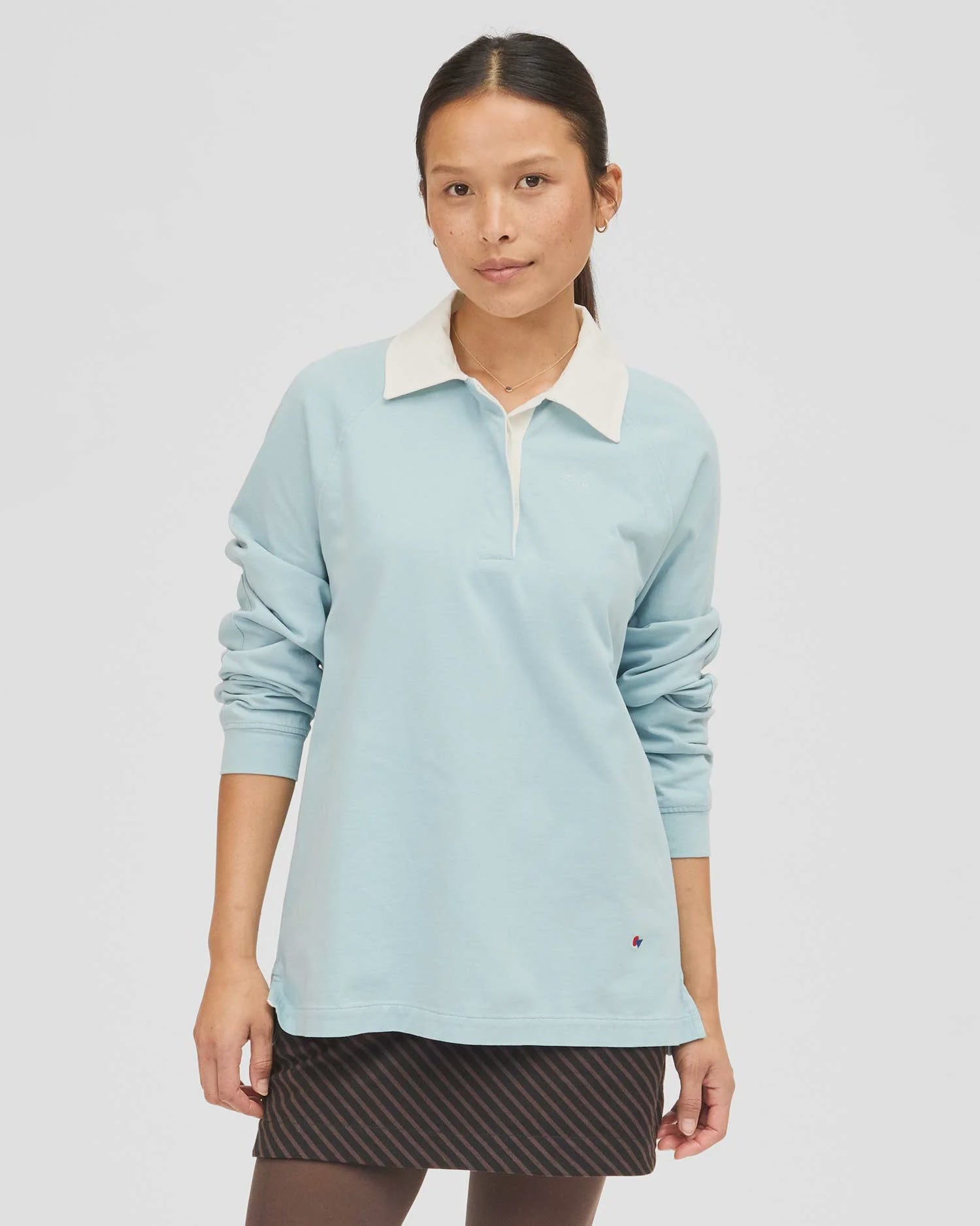 A person wearing Clare Vivier's Le Rugby, a light blue heavyweight cotton long-sleeved shirt with a white collar, stands against a white background. They pair it with a dark striped skirt with elbow patches, looking relaxed.