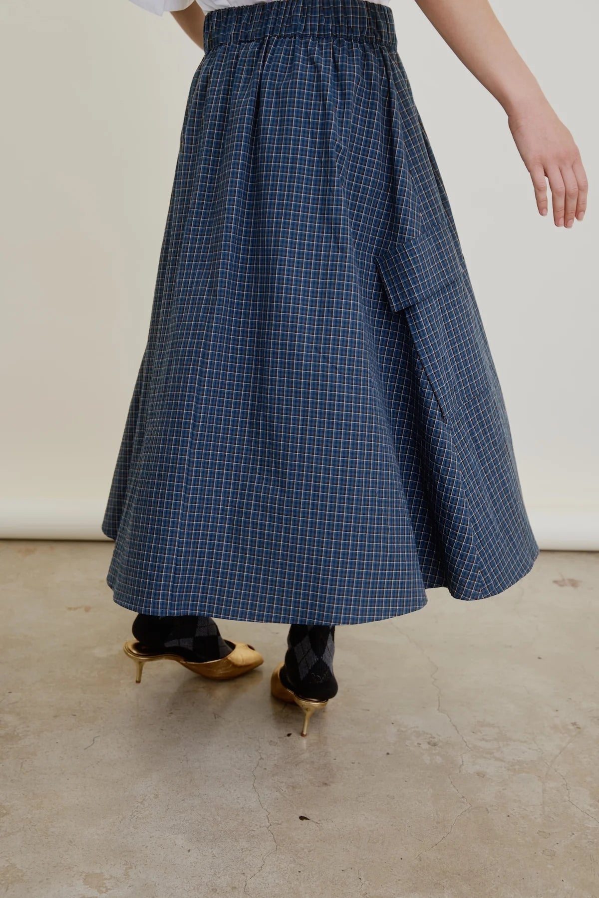 Wearing the Aquarius Cocktail Le Cargo Skirt—a long blue plaid cargo skirt with an elastic waist and pocket—this person pairs it with black patterned socks and gold heeled shoes while standing on a concrete floor.