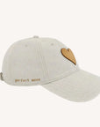 The KR Leather Heart Hat by Kerri Rosenthal is a light beige cotton-twill cap with a brown heart-shaped patch on the front, "perfect mess" embroidery on the side, and an adjustable fit for stylish comfort.