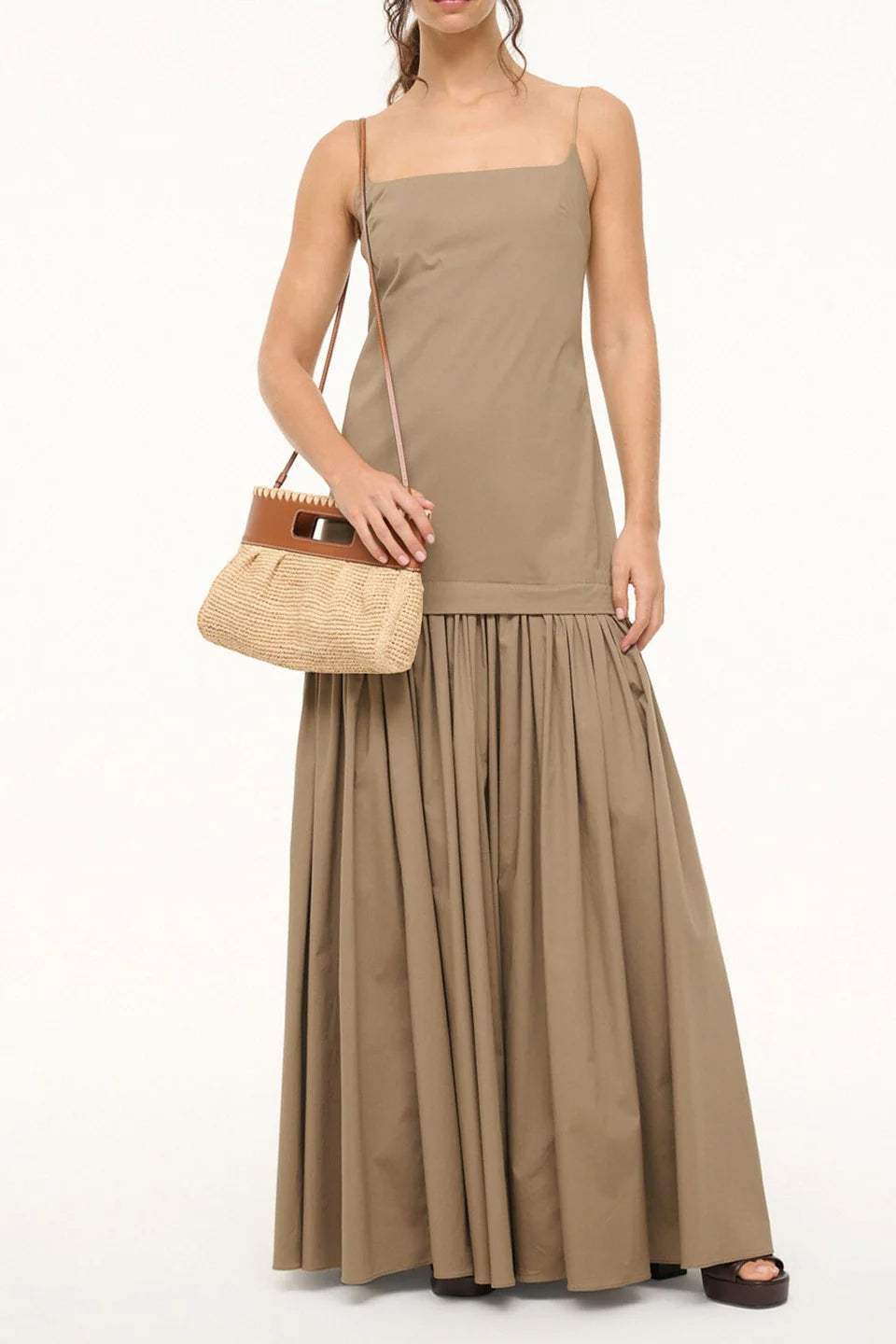 A woman wears a sleeveless tan maxi dress with a square neckline and gathered skirt. She carries the Lena Raffia Clutch by Staud and wears brown sandals. The background is plain white.