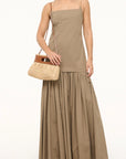 A woman wears a sleeveless tan maxi dress with a square neckline and gathered skirt. She carries the Lena Raffia Clutch by Staud and wears brown sandals. The background is plain white.