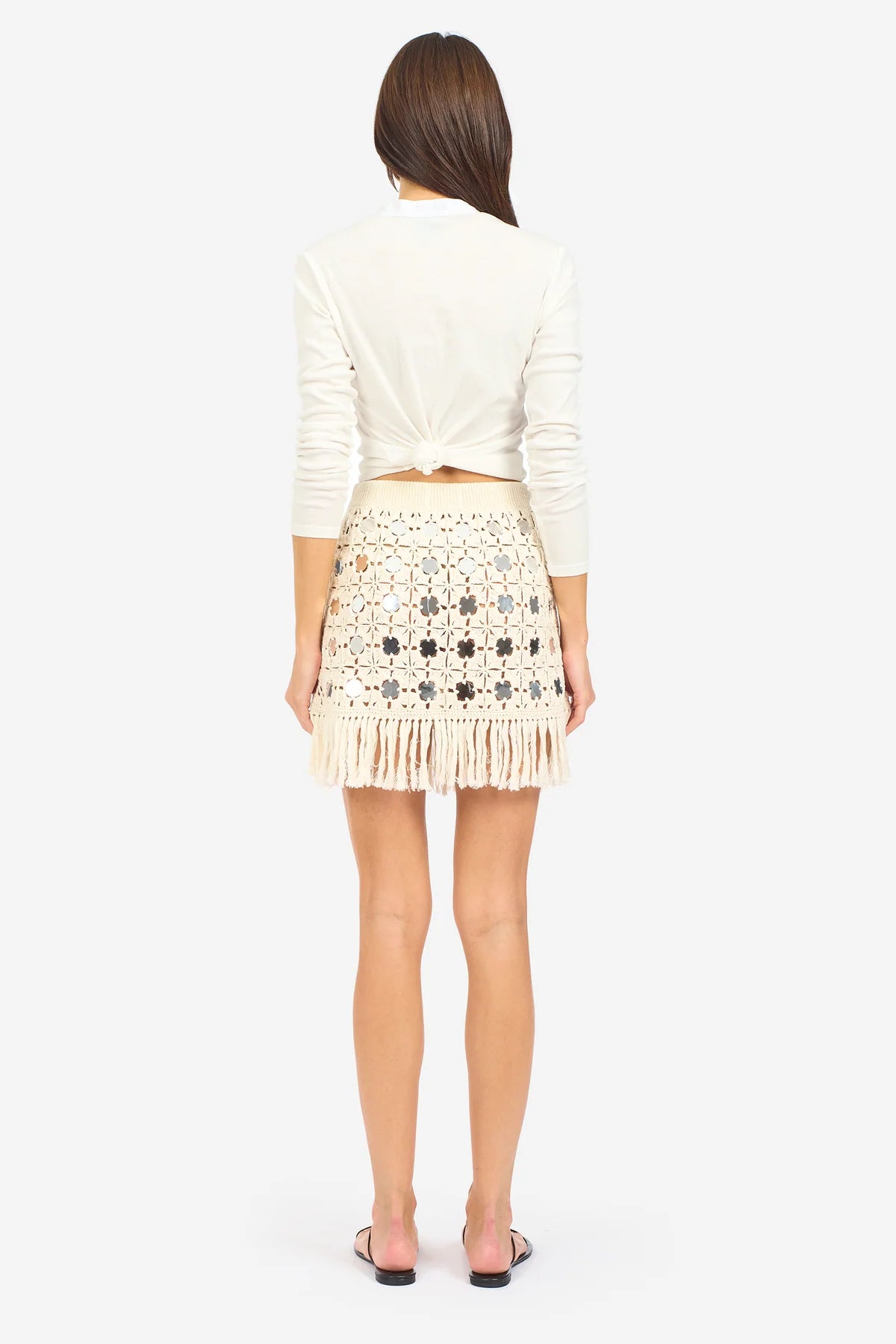 A woman with long brown hair stands with her back to the camera, wearing a white long-sleeve top and the Le Superbe Gypset Crochet Mirrorwed Mini Skirt, featuring cream crochet fabric and fringe at the hem. She pairs it with flat black sandals.