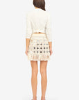 A woman with long brown hair stands with her back to the camera, wearing a white long-sleeve top and the Le Superbe Gypset Crochet Mirrorwed Mini Skirt, featuring cream crochet fabric and fringe at the hem. She pairs it with flat black sandals.