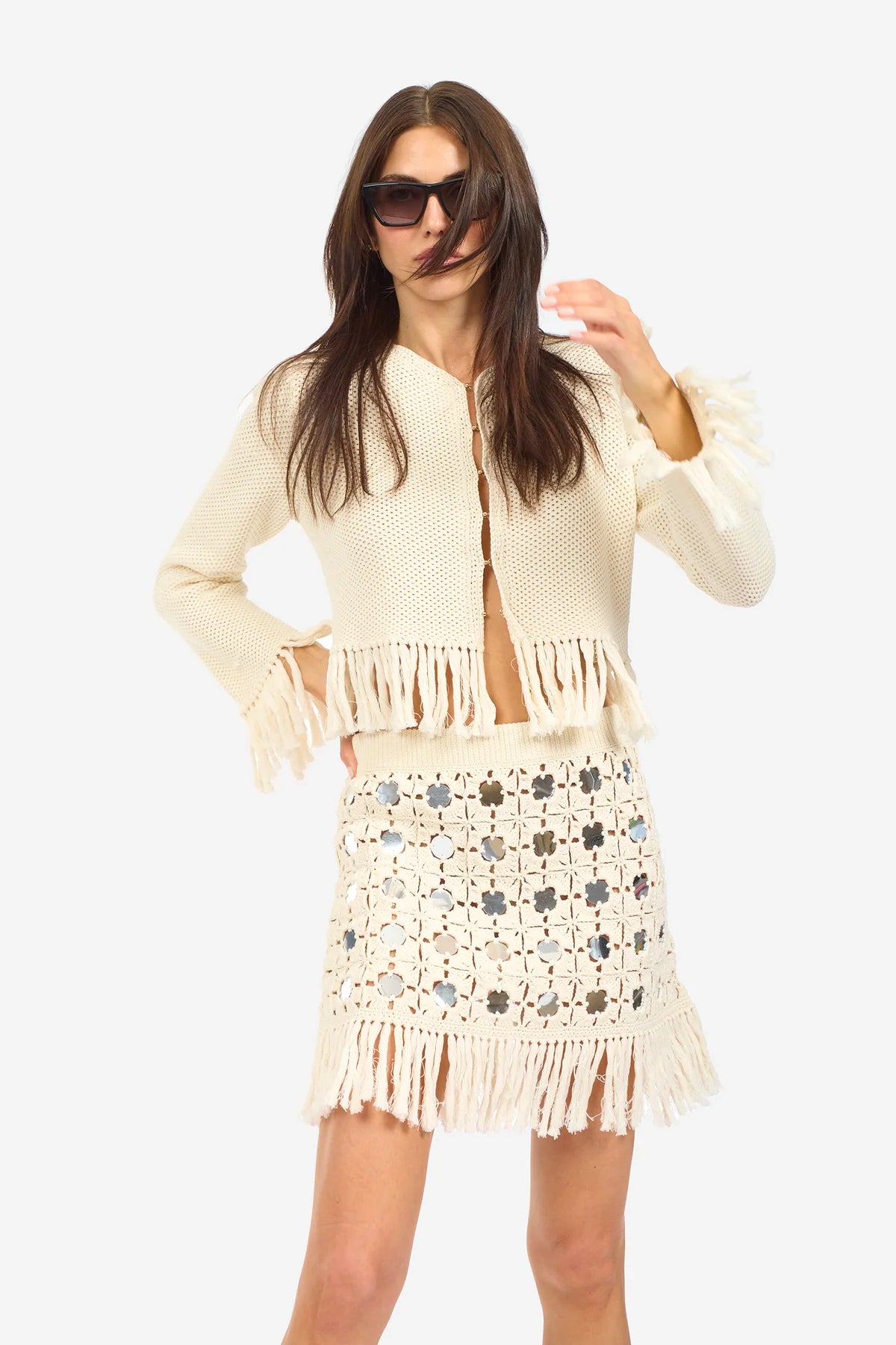 A woman wearing sunglasses, a cream knit jacket with fringe, and the Le Superbe Gypset Crochet Mirrorwed Mini Skirt poses against a plain white background.