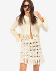 A woman wearing sunglasses, a cream knit jacket with fringe, and the Le Superbe Gypset Crochet Mirrorwed Mini Skirt poses against a plain white background.