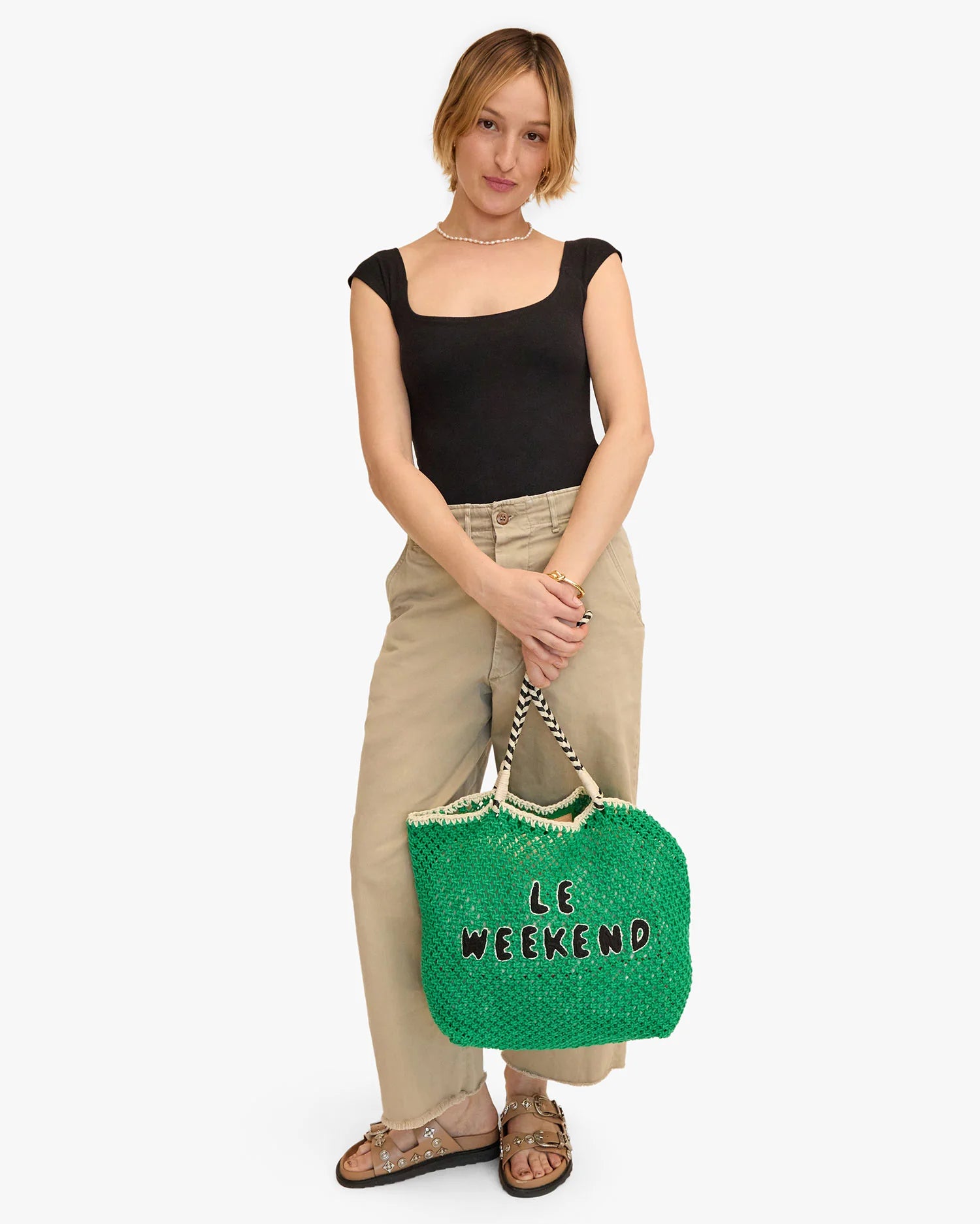 A person with short hair wears a black top, beige wide-legged pants, and sandals. They hold a large green Lete Tote by Clare Vivier with the words "Le Weekend" on it in black text. They have a neutral expression and stand in front of a plain white background.