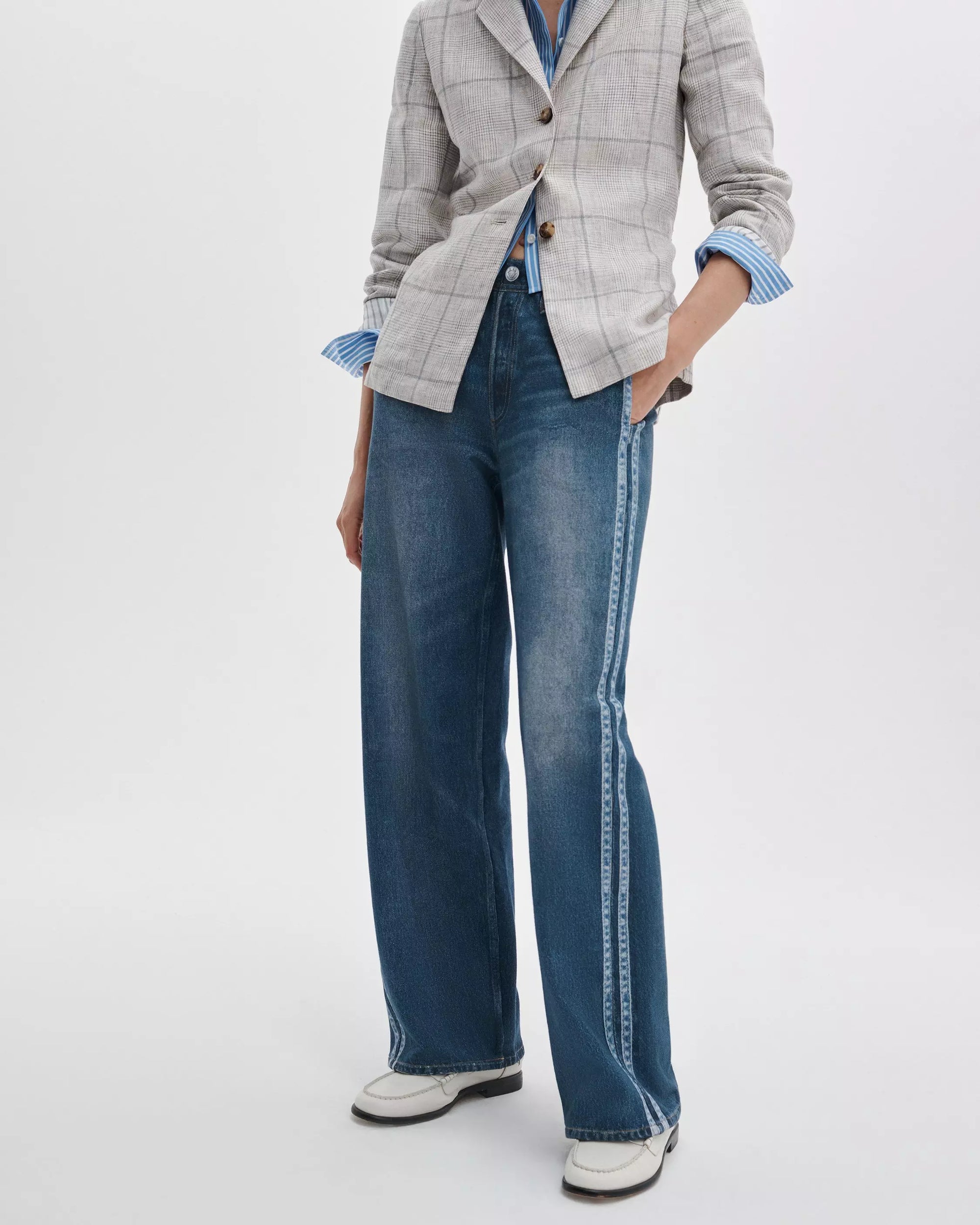 A person wearing a light gray plaid blazer, blue striped shirt, wide-leg blue jeans with side detailing, and Rag & Bone’s lexi loafers stands with one hand in their pocket against a plain background.