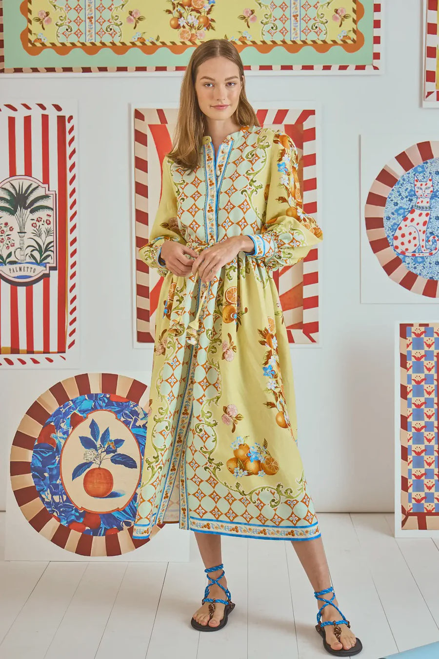 A woman models a Lillian Dress by Hunter Bell NYC, featuring a yellow floral pattern with blue accents, paired with black and blue strappy sandals. Colorful patterned artwork with stripes and florals adorns the crisp white wall backdrop.