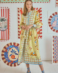 A woman models a Lillian Dress by Hunter Bell NYC, featuring a yellow floral pattern with blue accents, paired with black and blue strappy sandals. Colorful patterned artwork with stripes and florals adorns the crisp white wall backdrop.