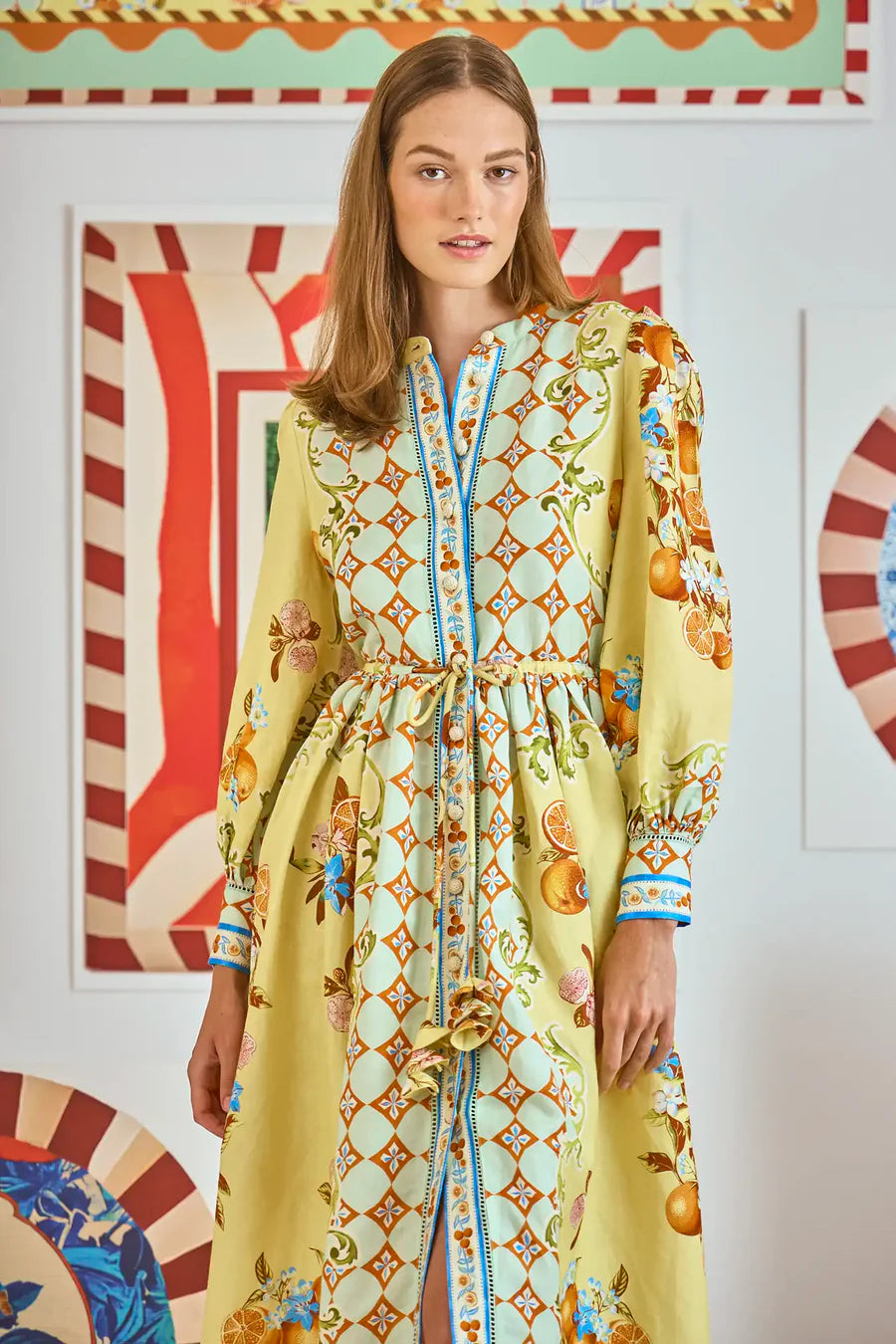 A woman in the Hunter Bell NYC Lillian Dress with geometric and floral patterns stands before colorful abstract artworks. Featuring a light green base with blue accents, it boasts a cinched waist and long sleeves, making it a timeless floral staple.